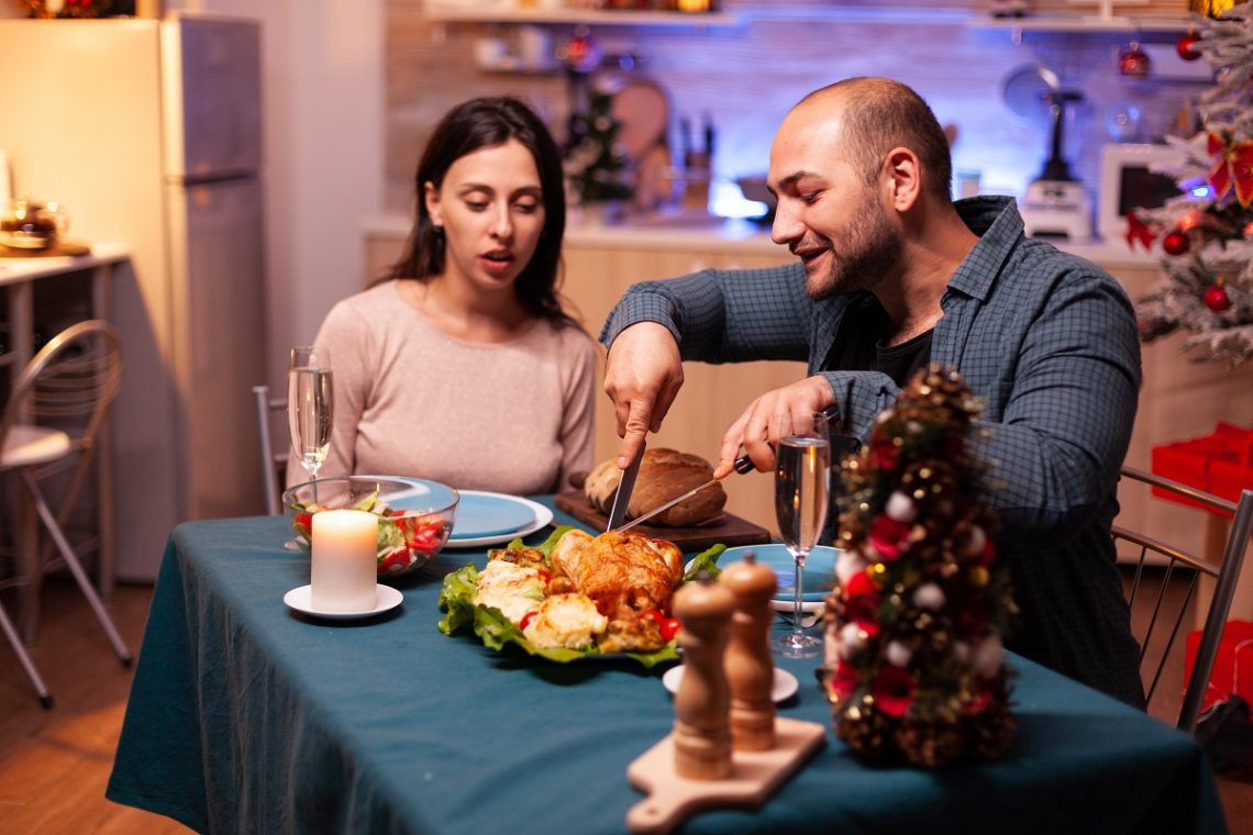 Happy family sitting at dining table in xmas decorated kitchen celebrating christmas holiday. Romantic cheerful couple eating xmas dinner enjoying spending winter season together